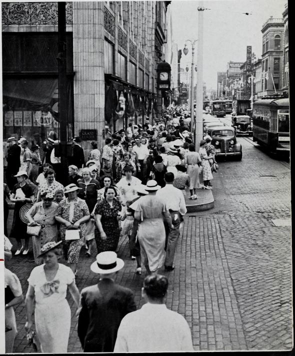 Large crowd on sidewalk crossing brick street at Grand Leader